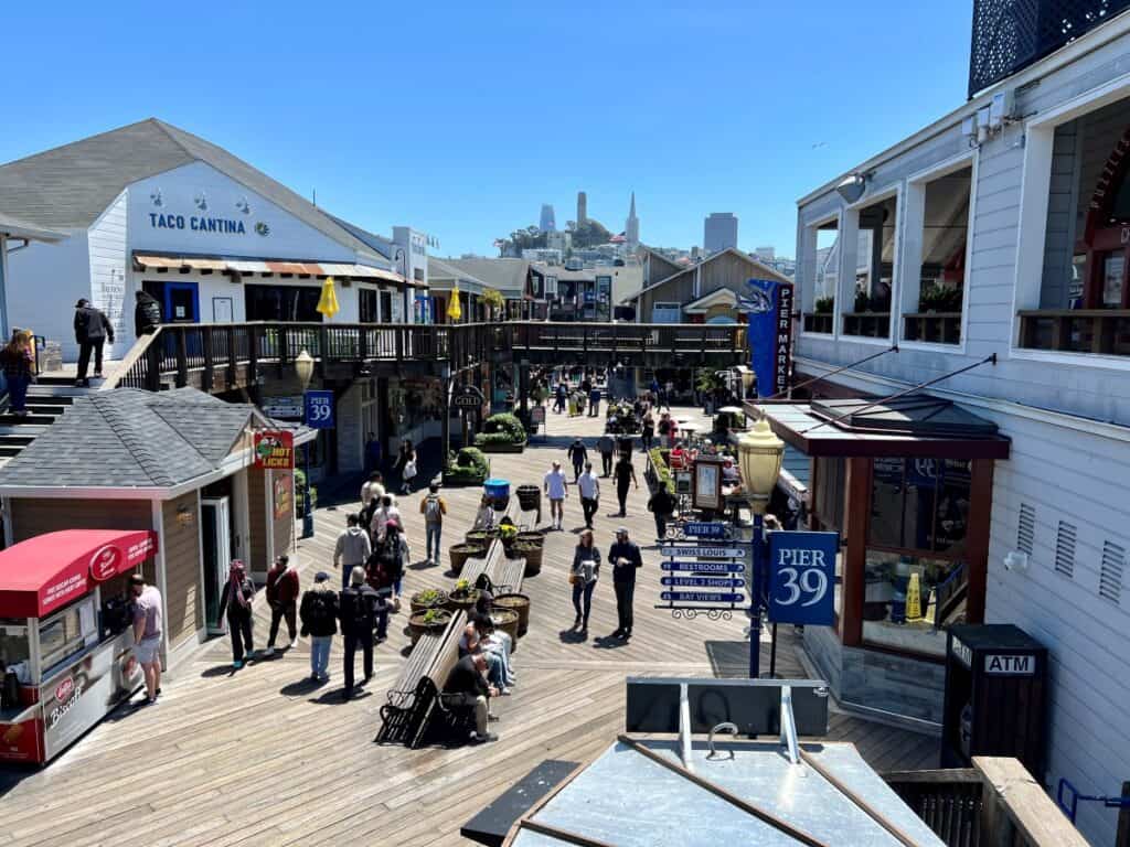 This image shows Pier 39 in San Francisco, featuring a lively scene with colorful shops and restaurants along the pier. The foreground includes tourists walking and enjoying the attractions, while the background displays the city skyline.