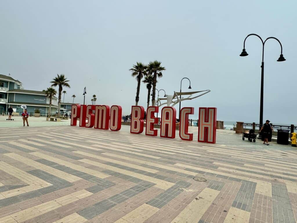 A tiled boardwalk in front of a beach with giant letters sign that reads Pismo Beach