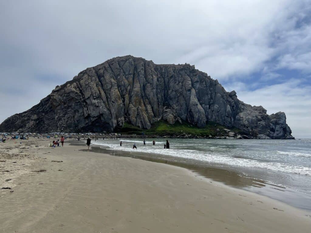 Morro Rock with a sandy beach and ocean waves in the foreground on a sunny day